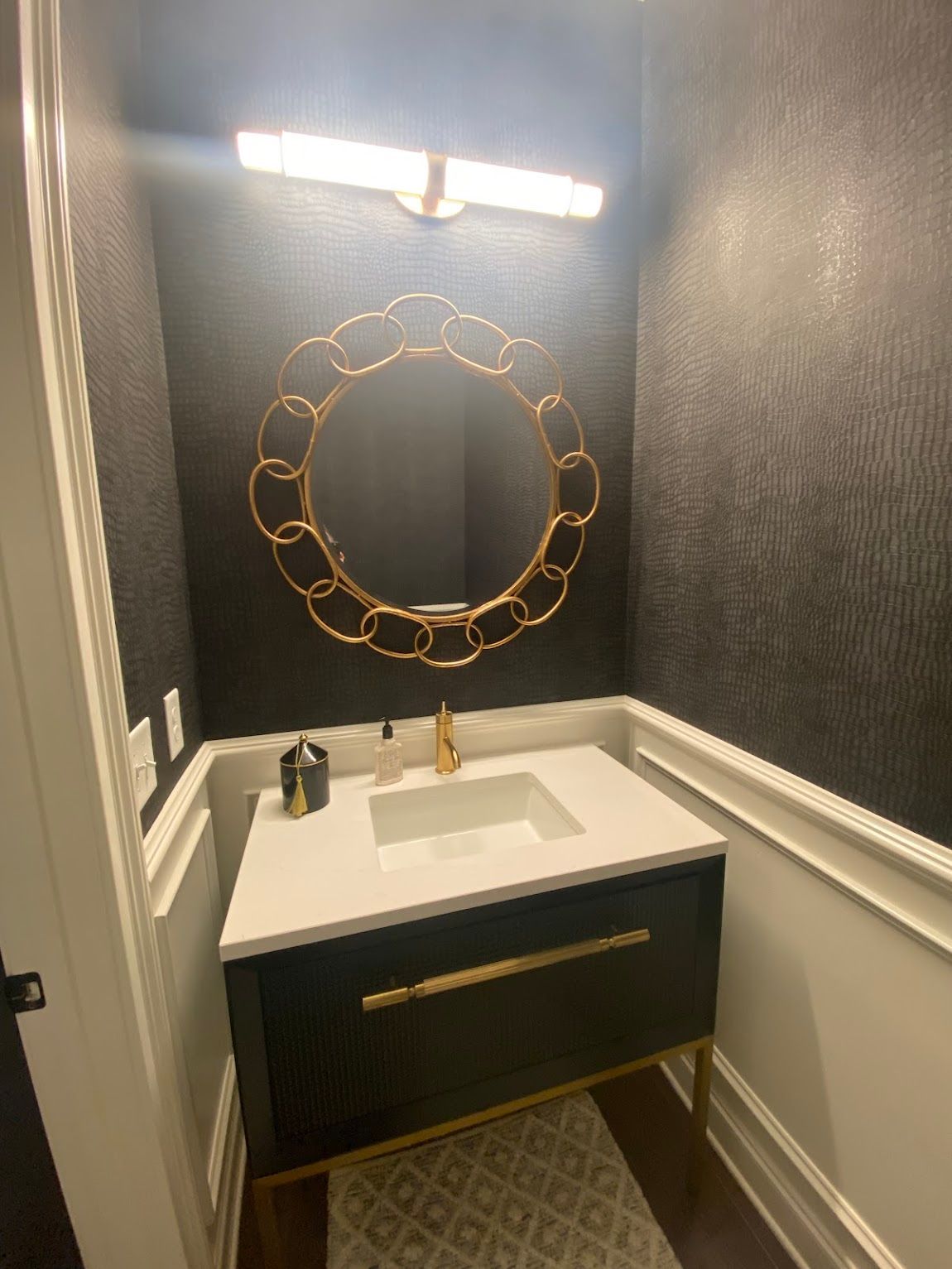 Dark powder room with gold accents: black vanity, round gold mirror, and modern lighting.