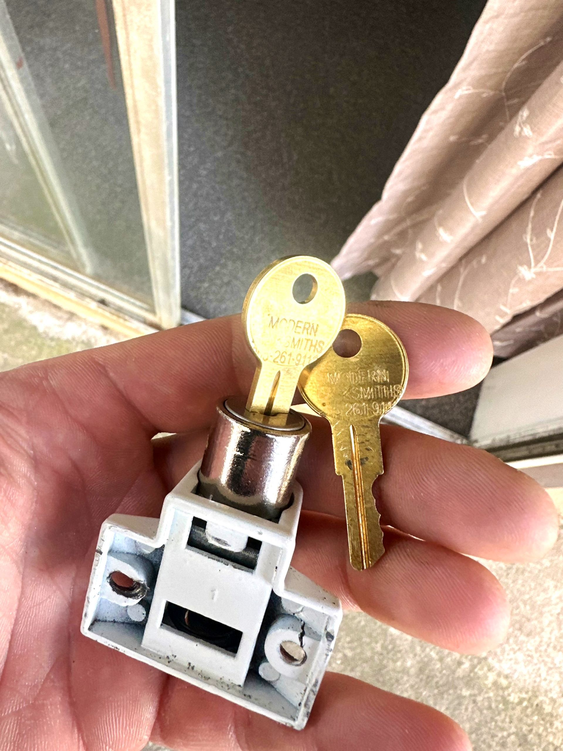 Hand holding a white window lock with two brass keys.