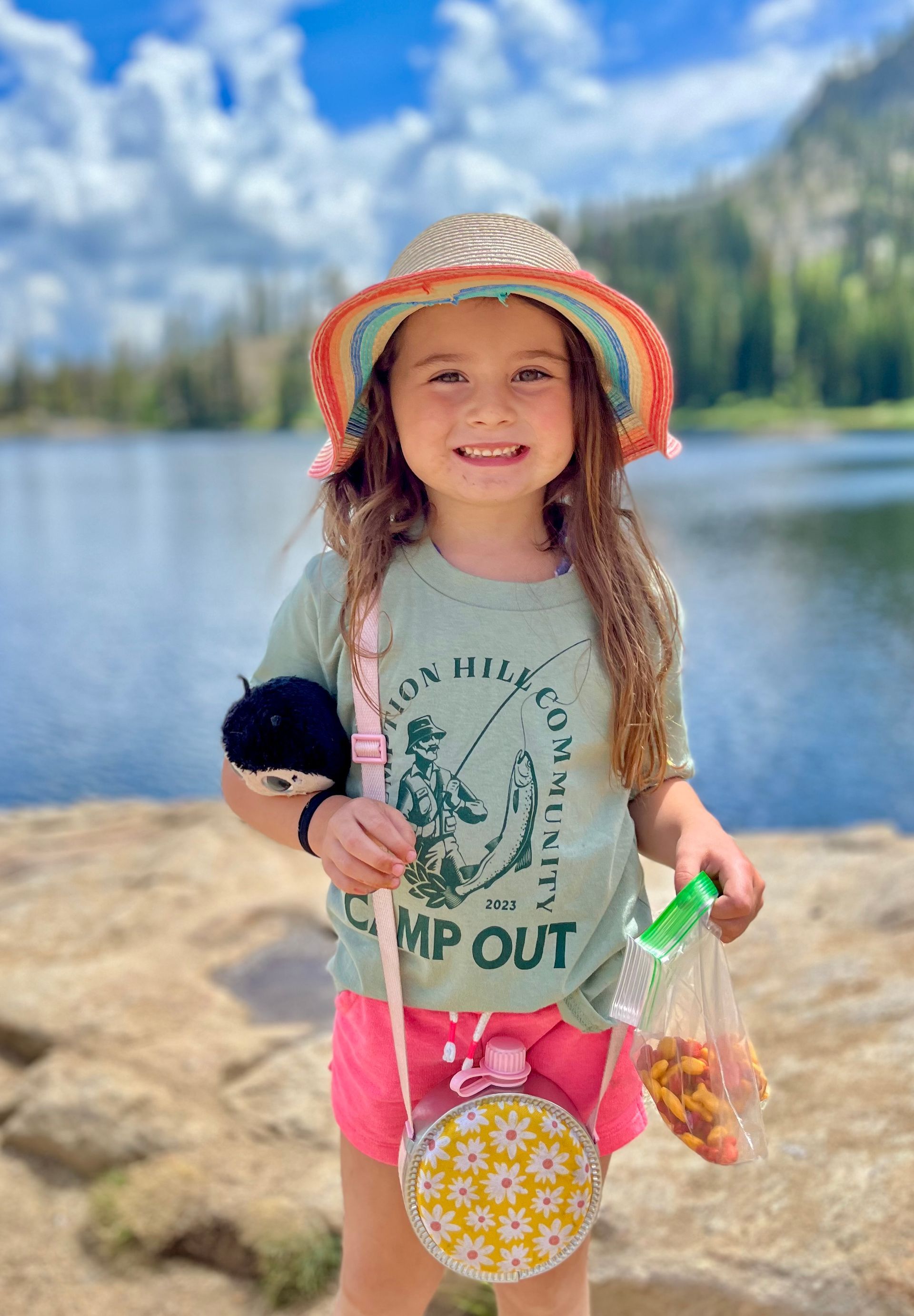 A little girl wearing a hat and a t-shirt is standing next to a lake.