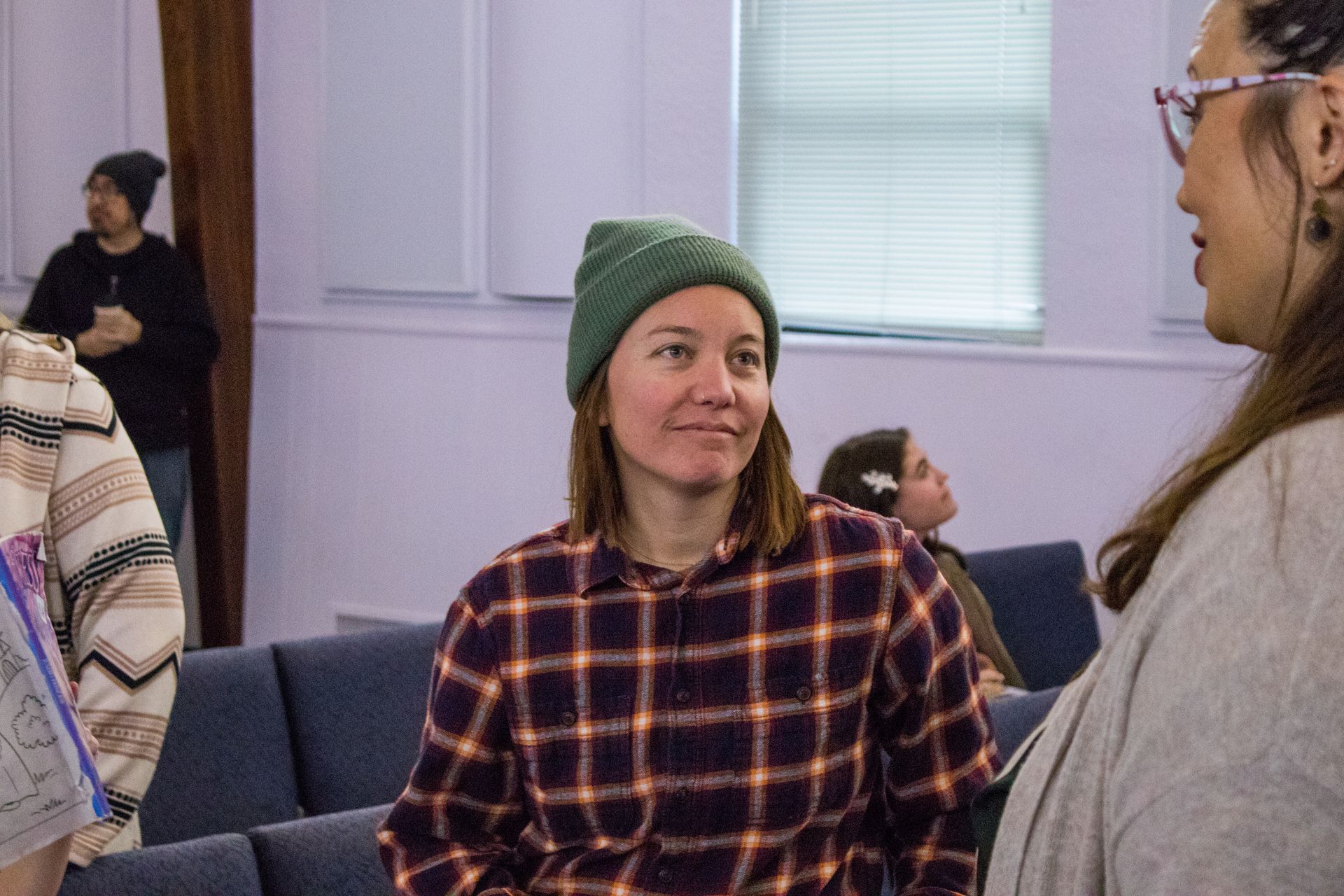 A woman wearing a plaid shirt and a green beanie is talking to another woman in a room.