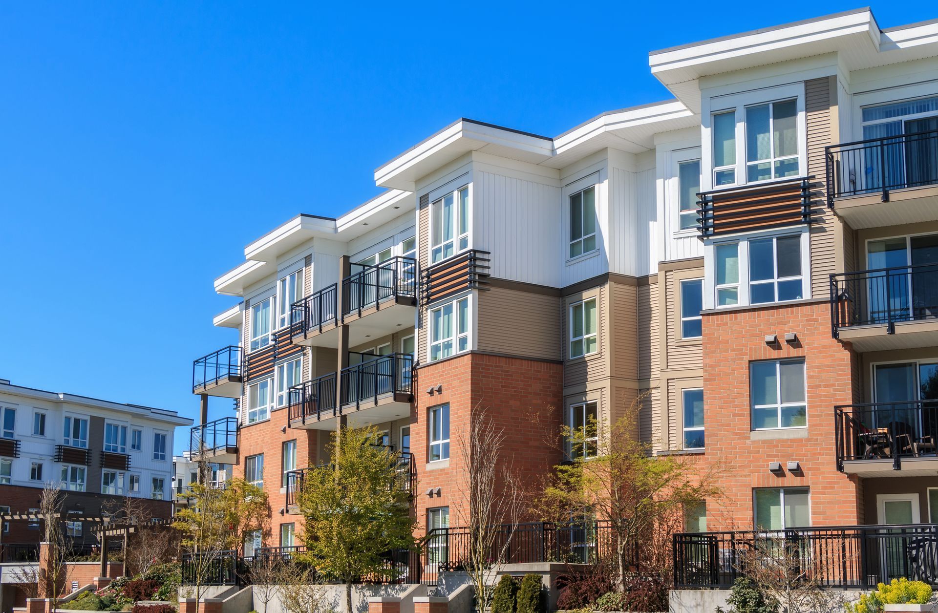Apartment building with red brick and white accents against a bright blue sky. Balconies and trees visible.