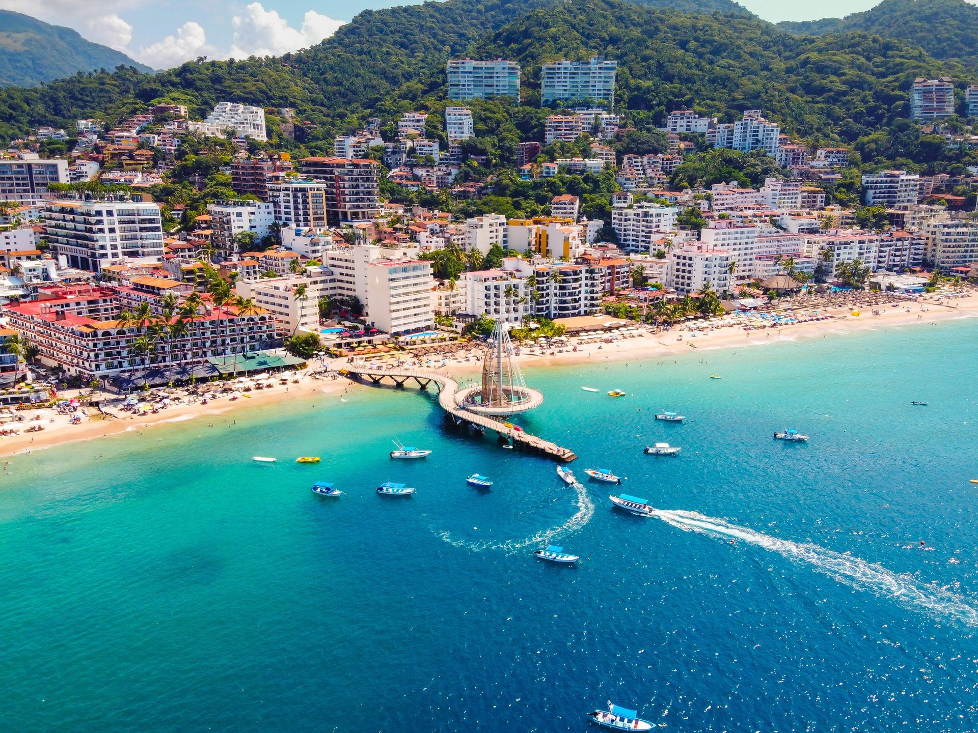 Aerial view of Puerto Vallarta, Mexico. Beach lined with buildings, boats on blue water, mountains in the background.