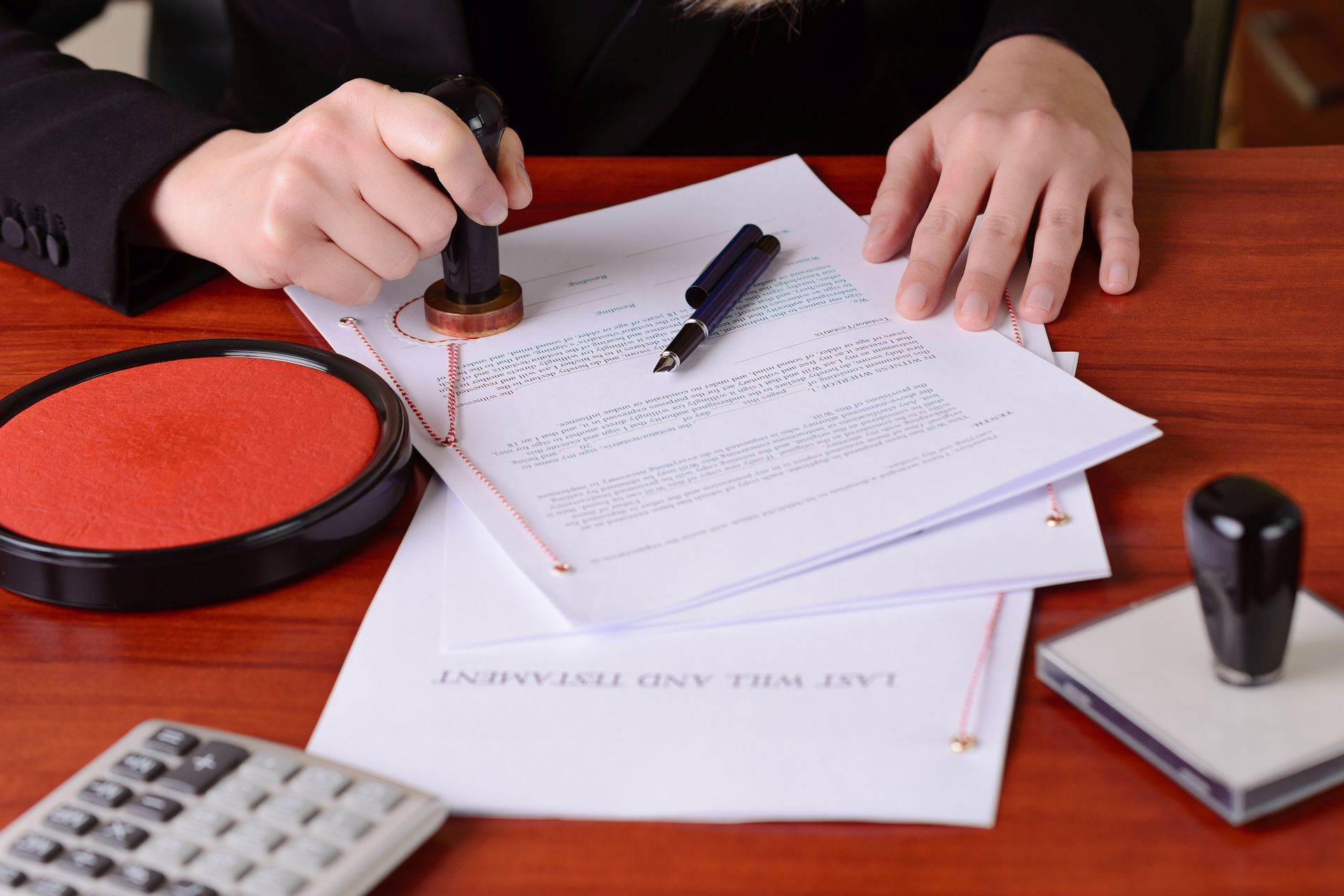 A person stamps a document, presumably a will, on a desk, near a red ink pad, a pen, and a calculator.