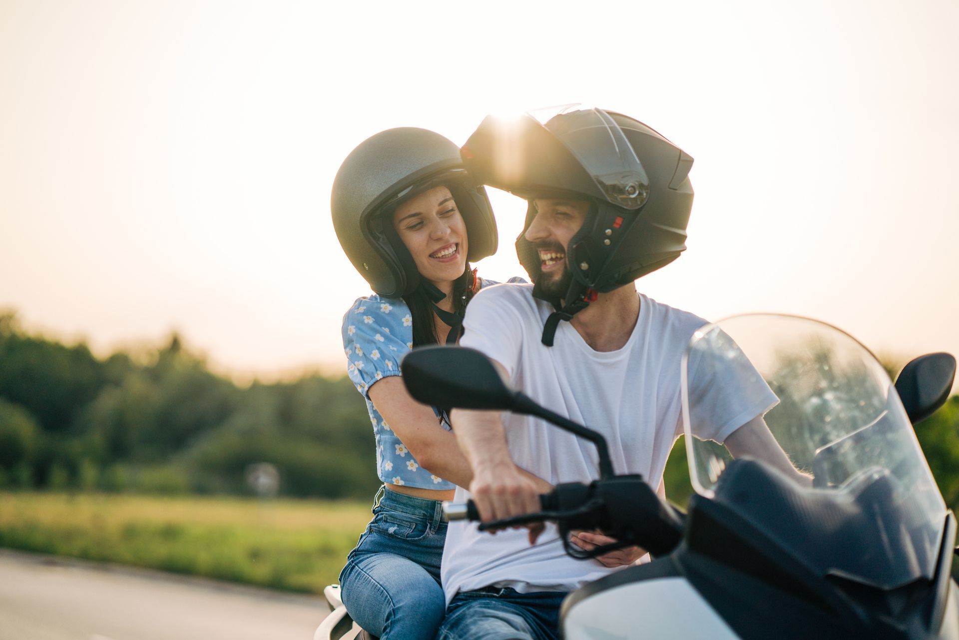 A couple on a scooter, wearing helmets, driving on a road in the sunlight. The woman leans on the man with an endearing smile.