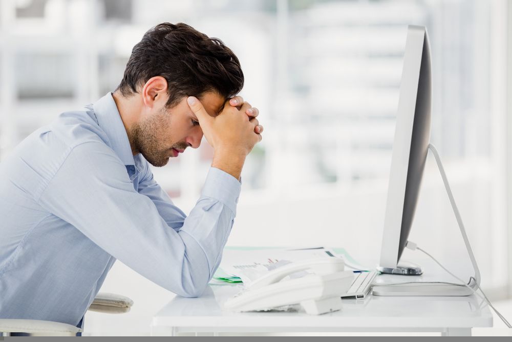 A man is sitting at a desk with his head in his hands in front of a computer.