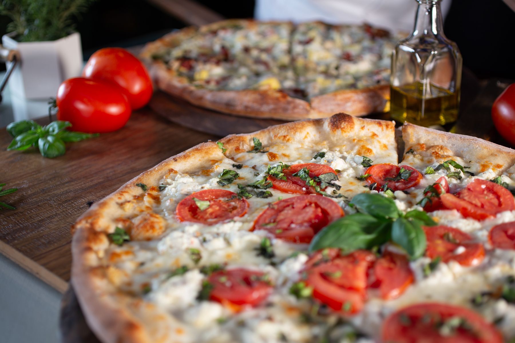 Two pizzas are sitting on a wooden table with tomatoes and basil.