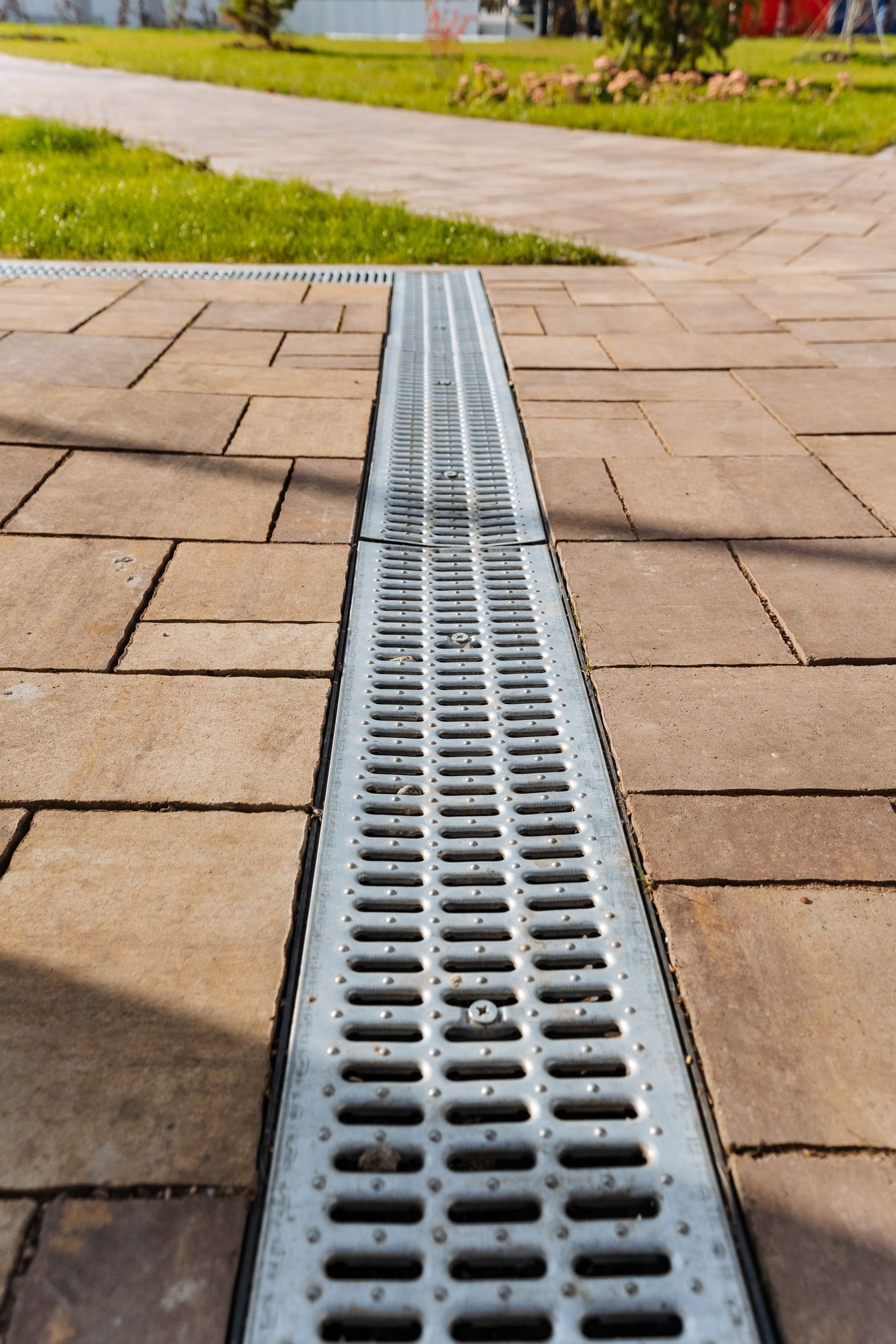 A long, metal grate drain in a paved walkway next to grass and a path.