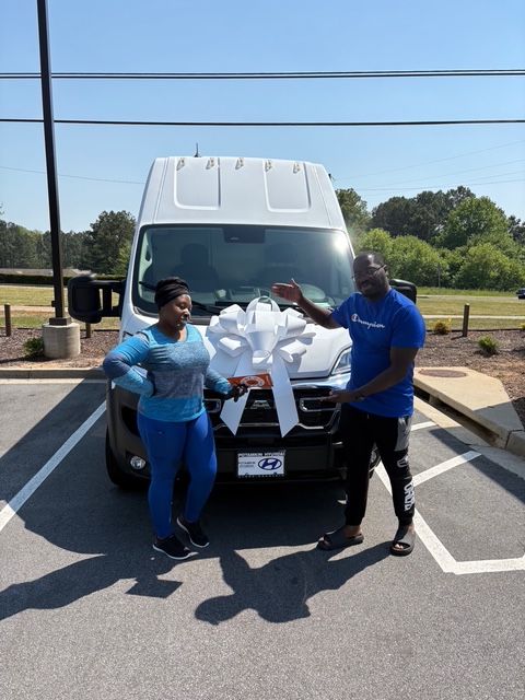 Woman and man pose in front of a white van with a bow on it, under a bright, sunny sky.