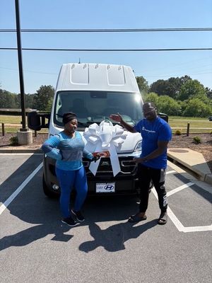 Woman and man pose in front of a white van with a bow on it, under a bright, sunny sky. Woman and man pose in front of a white van with a bow on it, under a bright, sunny sky.