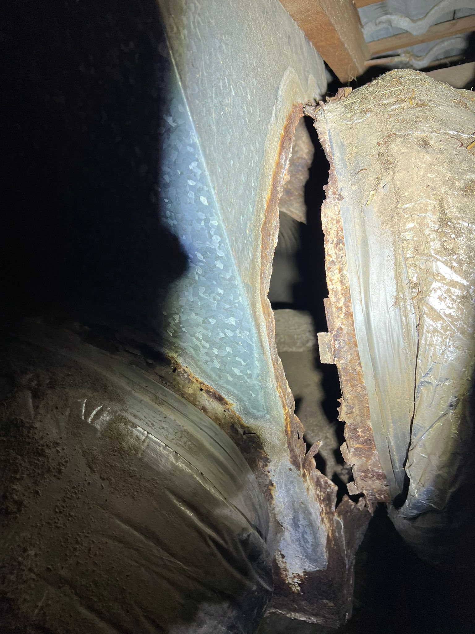 A dark, close-up view of severely rusted metal ductwork with a large, jagged hole in a dimly lit crawlspace.