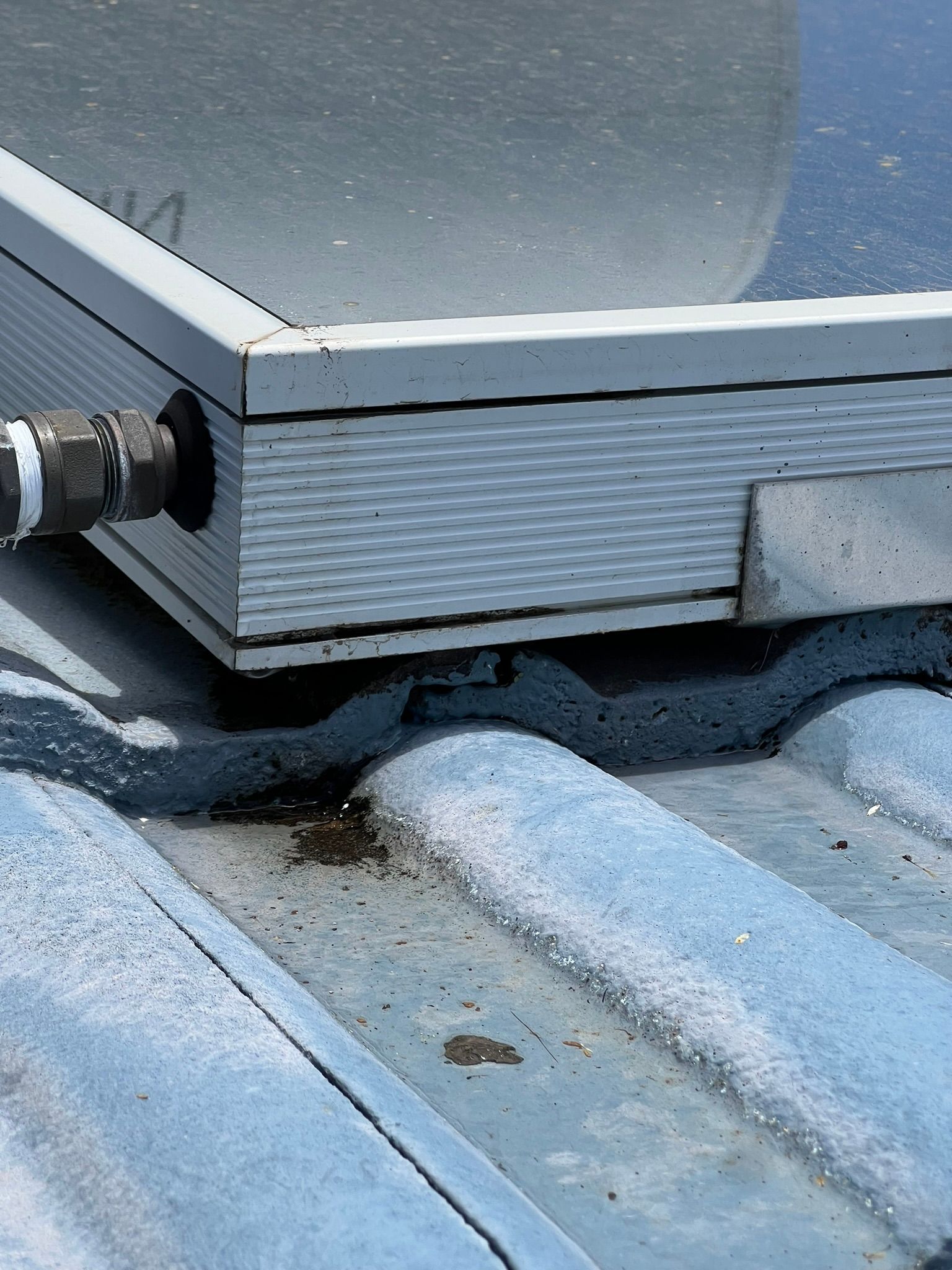 A close-up view of a metal solar panel frame resting on a corrugated grey roof, featuring a pipe fitting on the left.