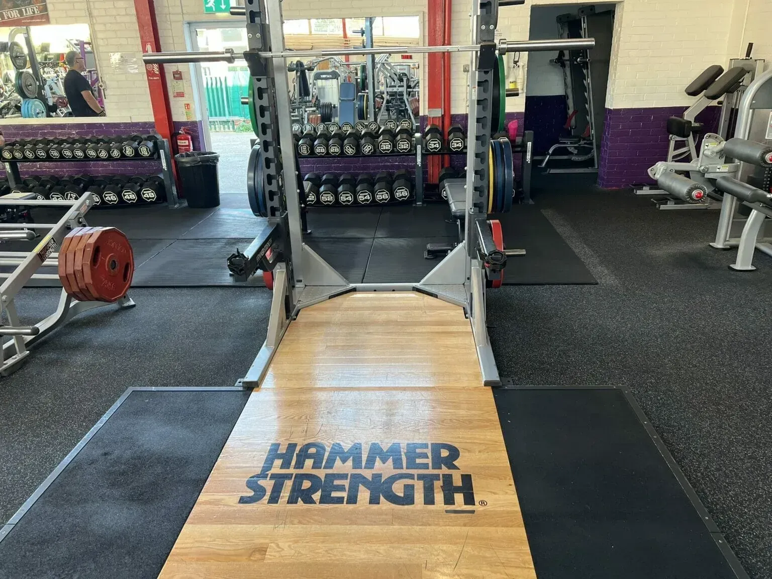 A Hammer Strength squat rack and weightlifting platform in a gym, with barbells and racks of weight plates in the background.