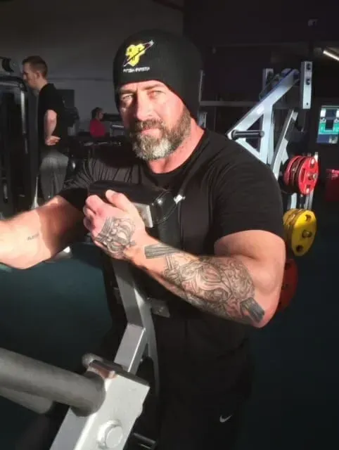 A bearded person in a gym, wearing a black beanie and t-shirt, working out on a resistance machine.