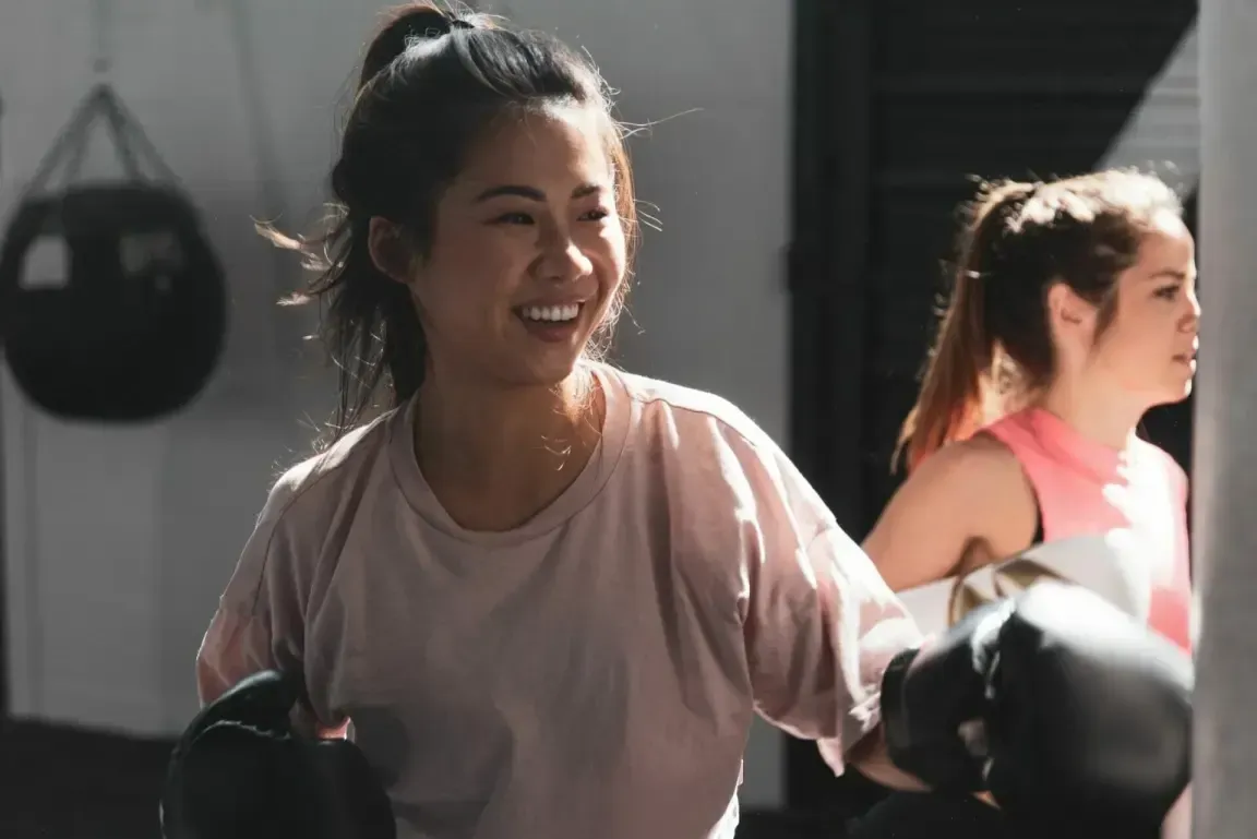 Two people with high ponytails wearing boxing gloves and athletic wear, smiling while hitting a punching bag at a gym.