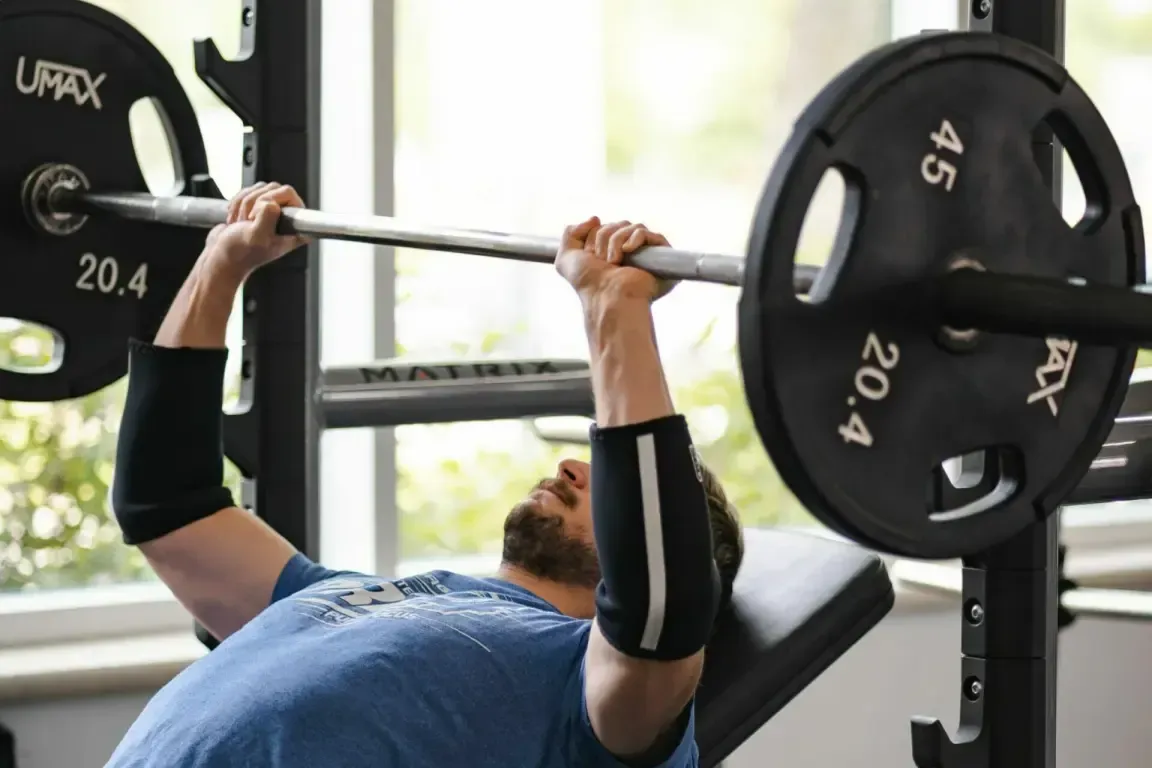 A person performs an incline barbell bench press at a gym while wearing black elbow sleeves.