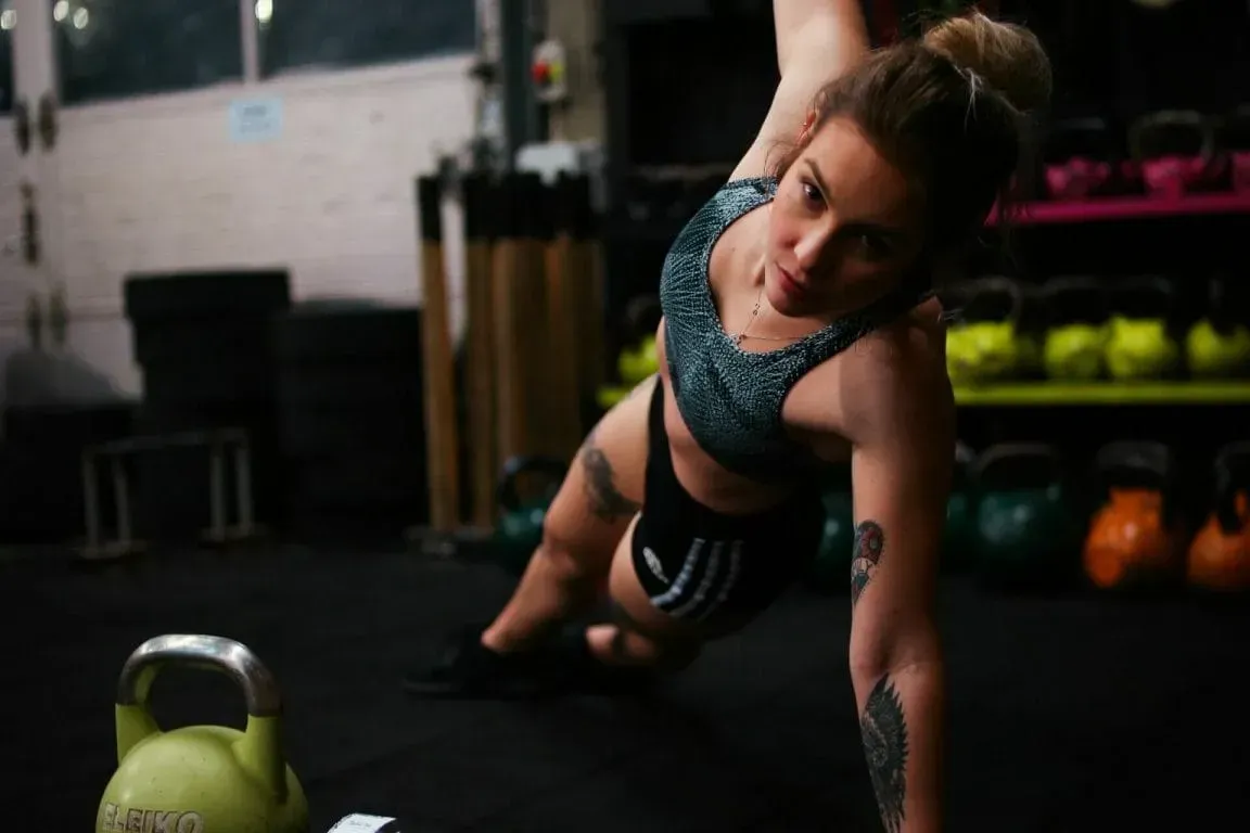A person holding a side plank position on a gym floor with kettlebells on shelves in the background.