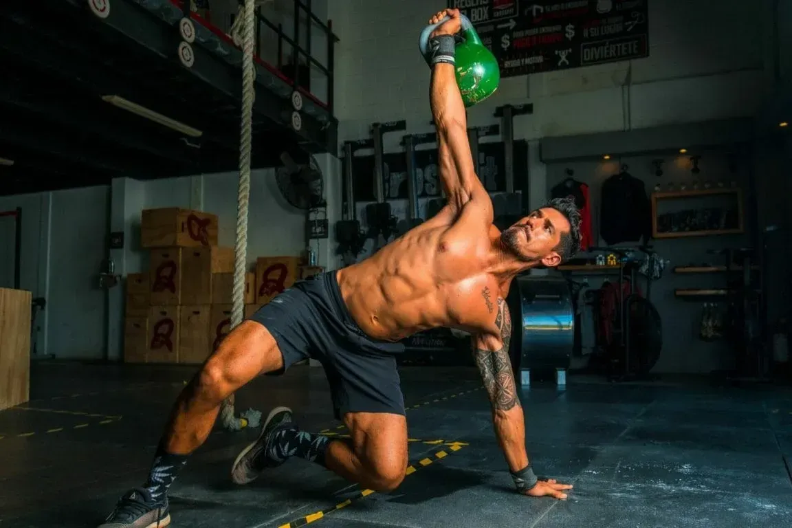 A shirtless person in shorts performs a kettlebell Turkish get-up in a gym.