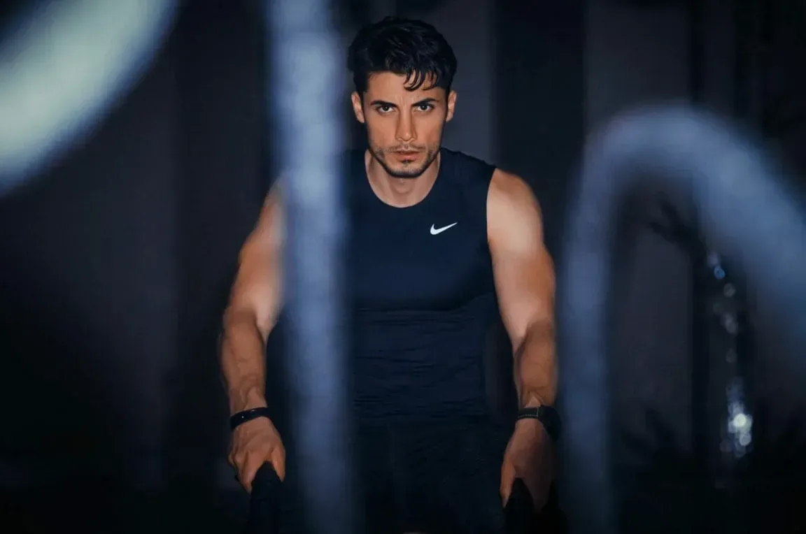 A focused individual wearing a black Nike tank top exercises with battle ropes in a dimly lit, blue-toned gym setting.