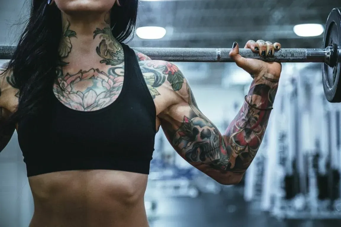 A person with tattooed arms and torso holds a barbell across their shoulders while working out in a gym.