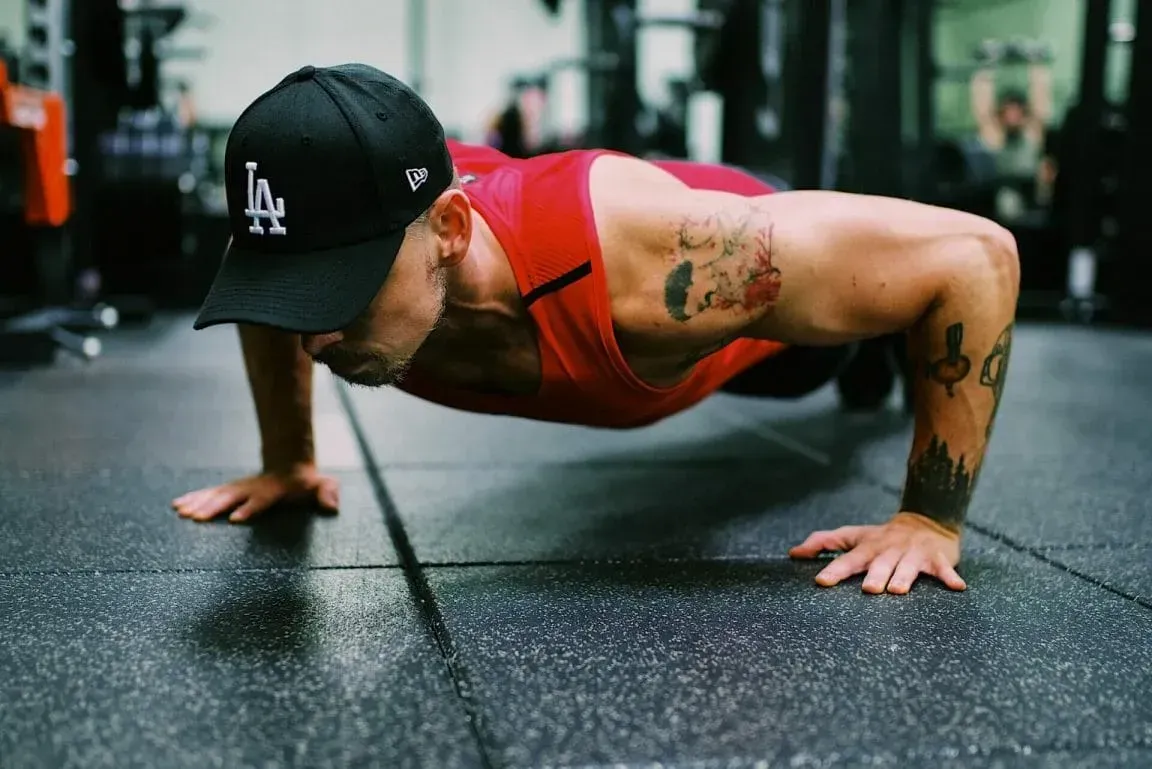 A person with tattoos wears a red tank top and black LA baseball cap while doing a push-up on a gym floor.