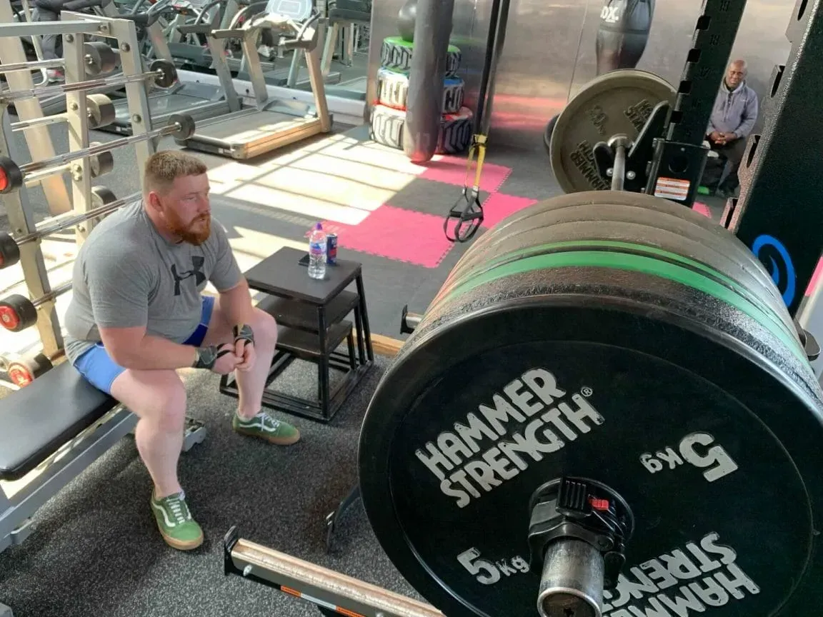 A person sits on a bench in a gym next to a barbell loaded with Hammer Strength weight plates.
