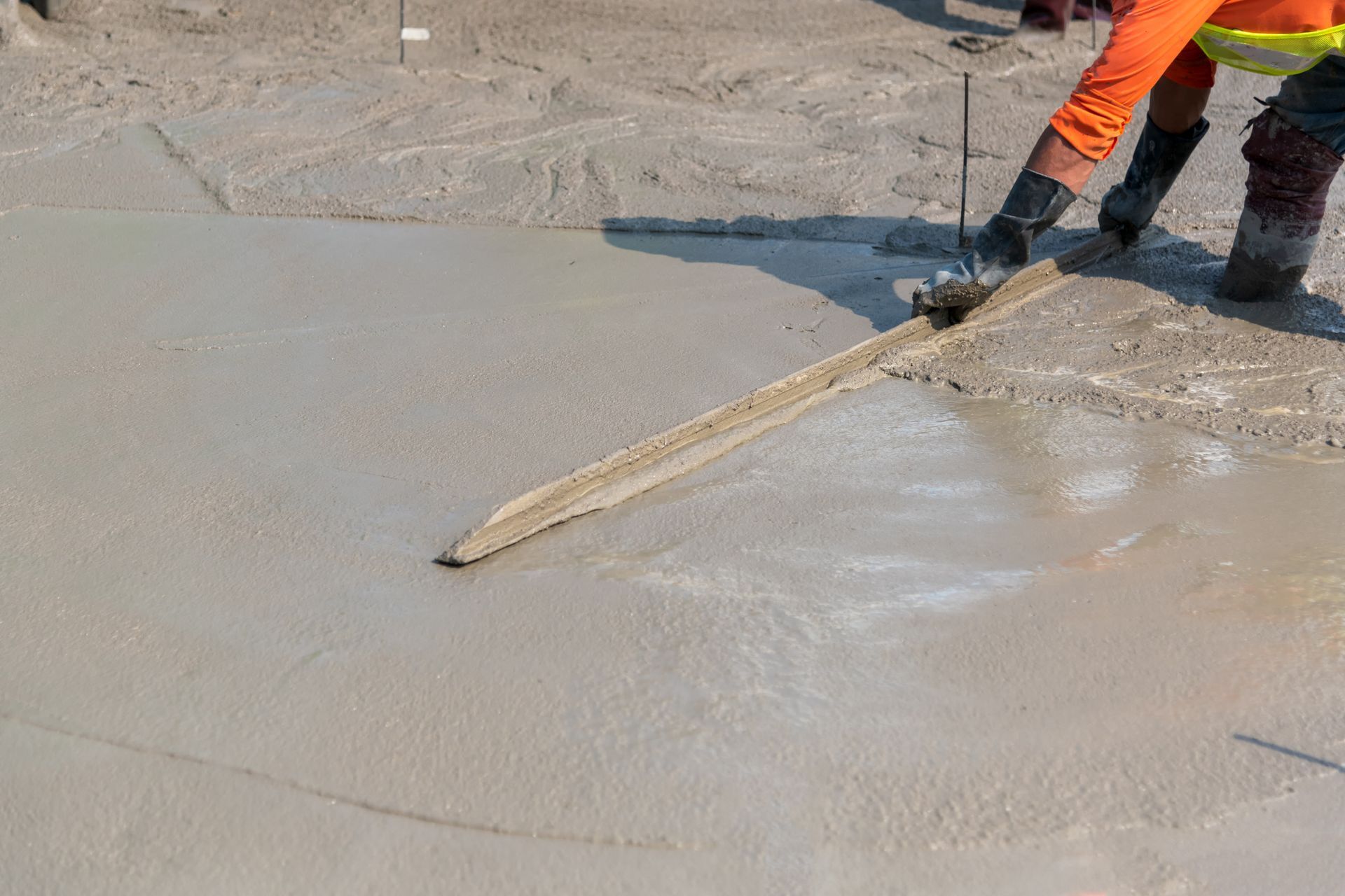 A construction worker is spreading concrete on the ground with a trowel.