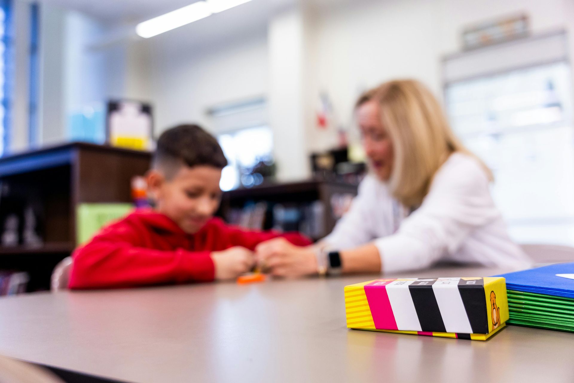 Child and teacher at a table, working on a project. A colorful block is in the foreground.