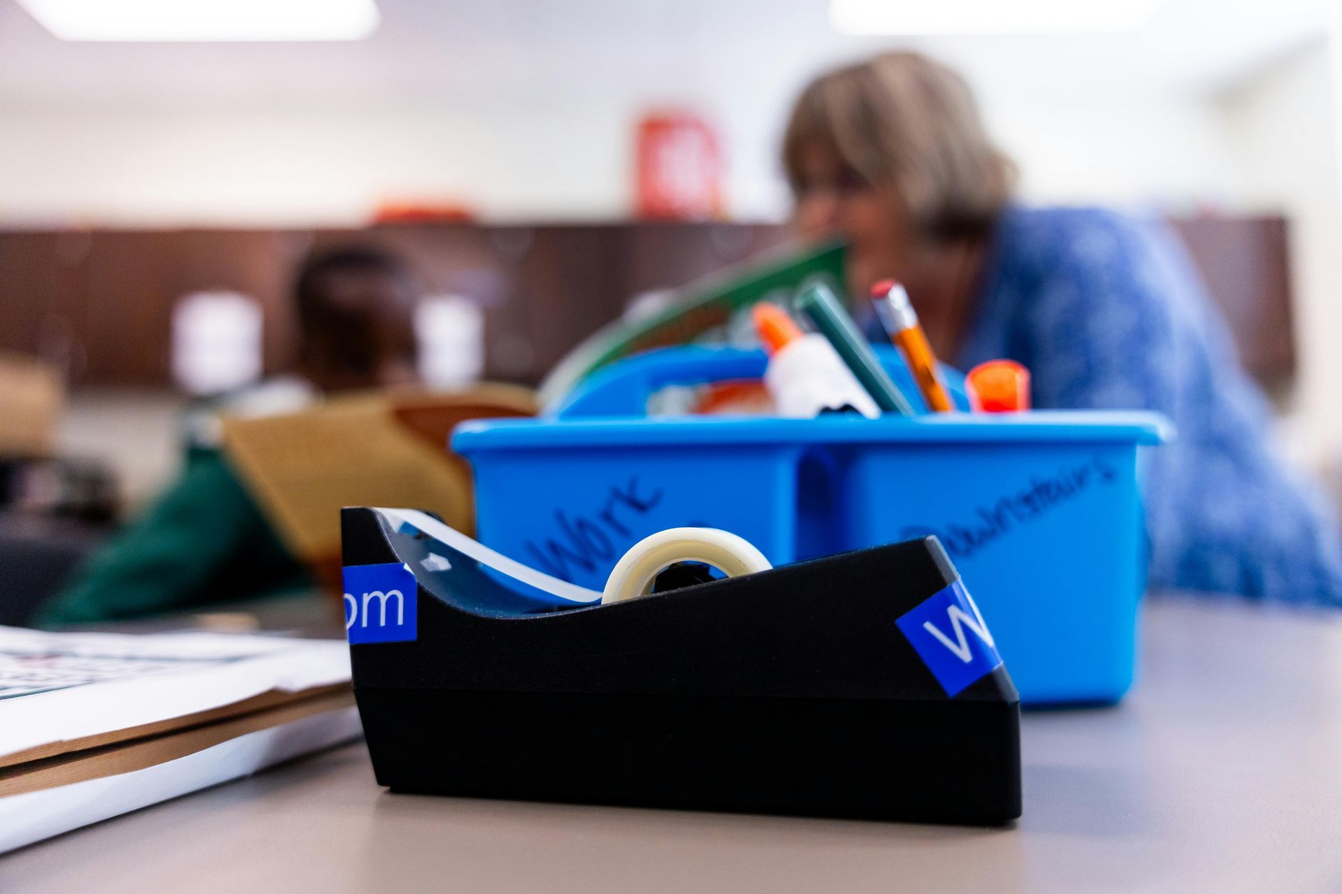 Black tape dispenser in focus on table; blue toolbox with supplies, and people in background.