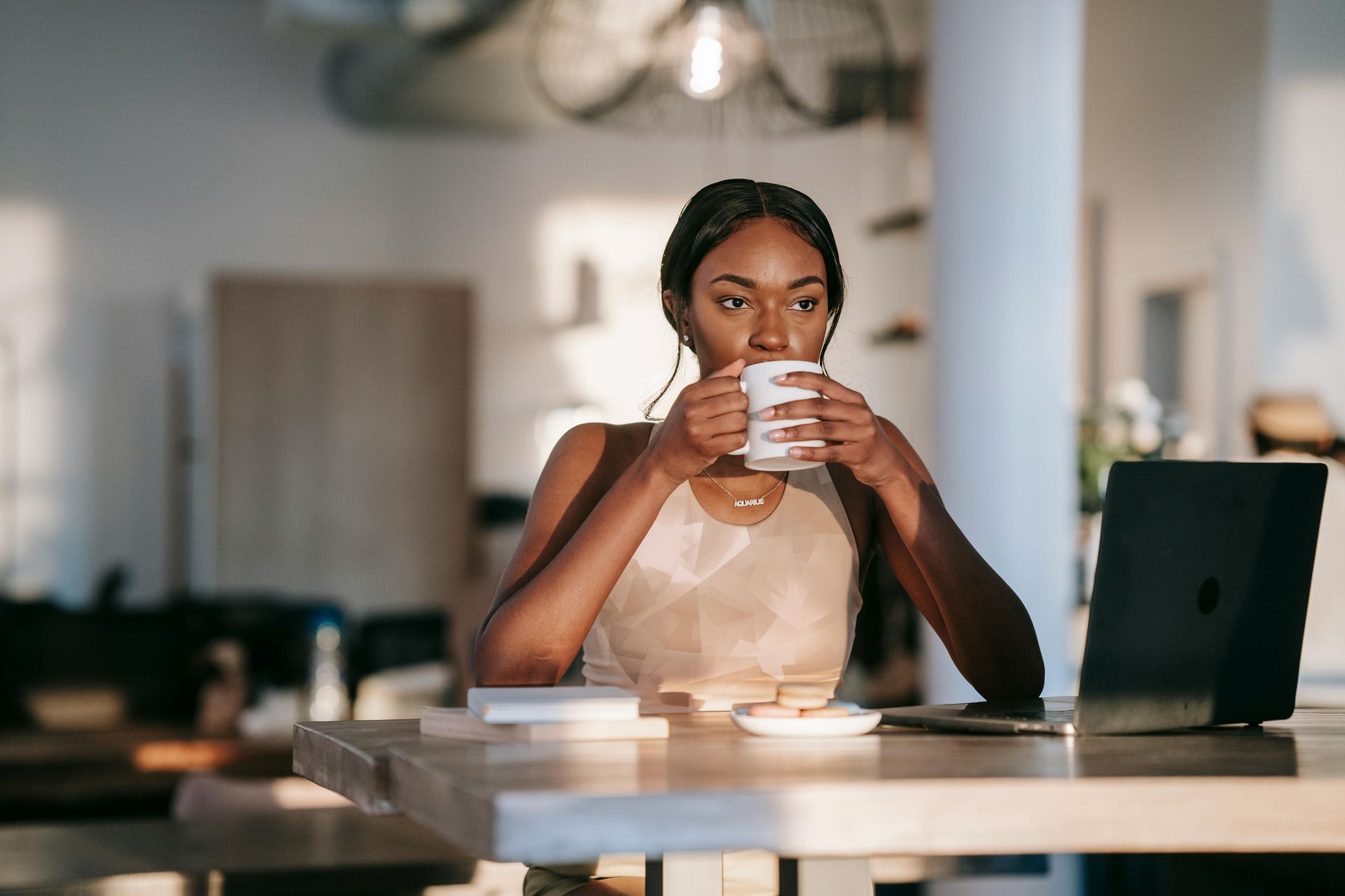 Woman sipping from a mug, working on a laptop at a wooden table in a bright room.