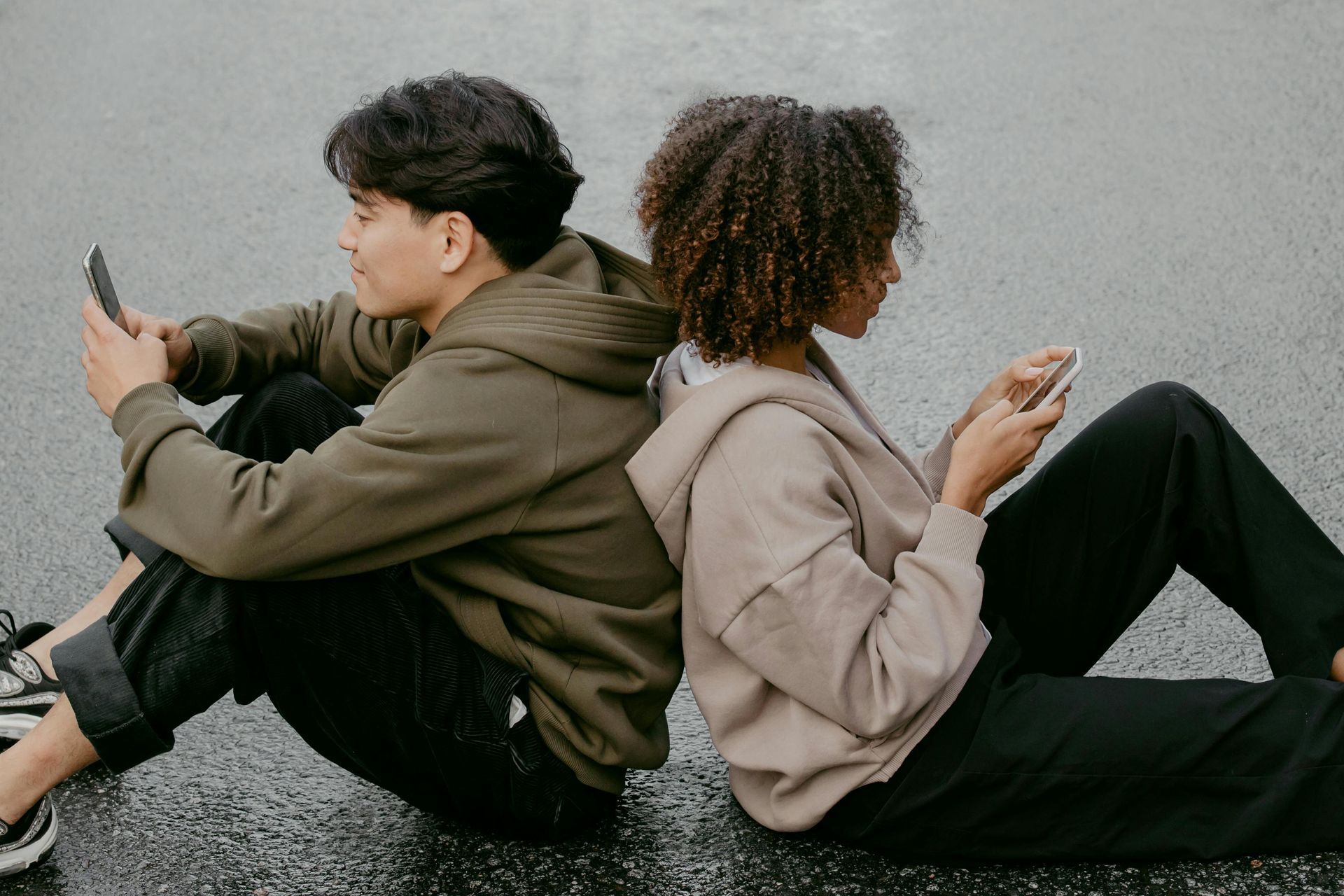 A man and a woman are sitting back to back on the ground looking at their phones.