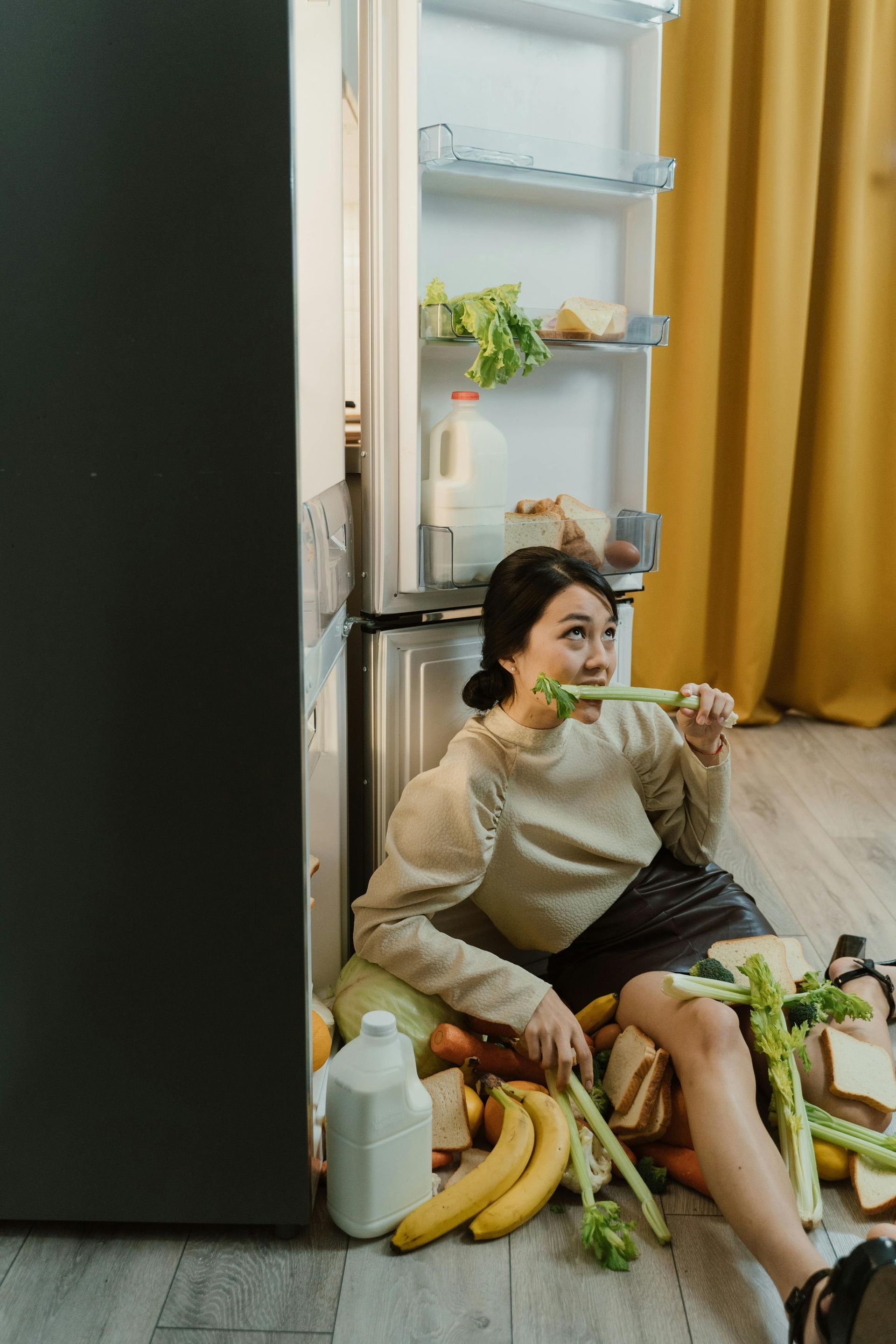 A woman is sitting on the floor in front of an open refrigerator eating celery.