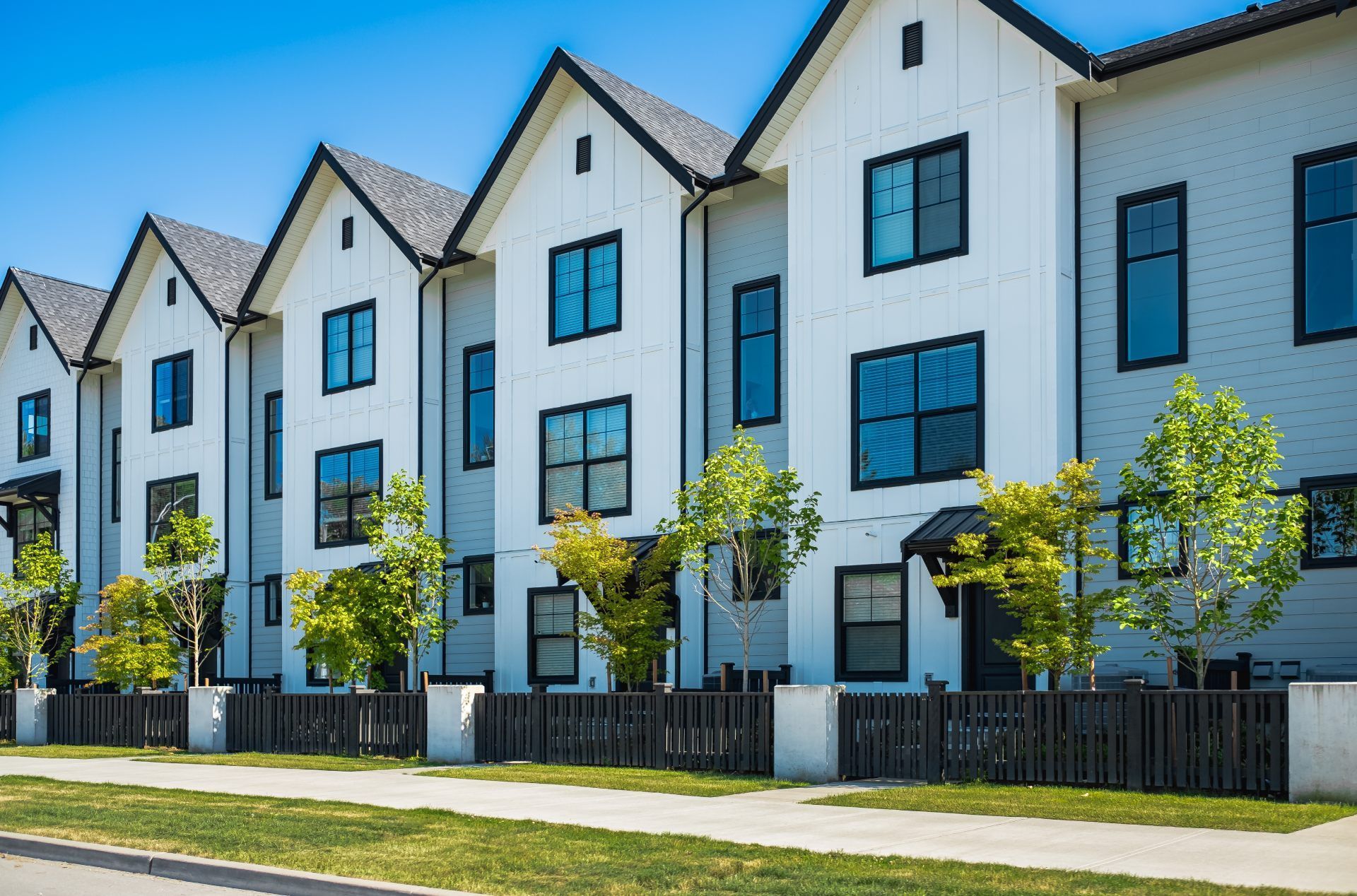 Row of modern, white townhouses with black trim and dark roofs; trees and a fence in front.