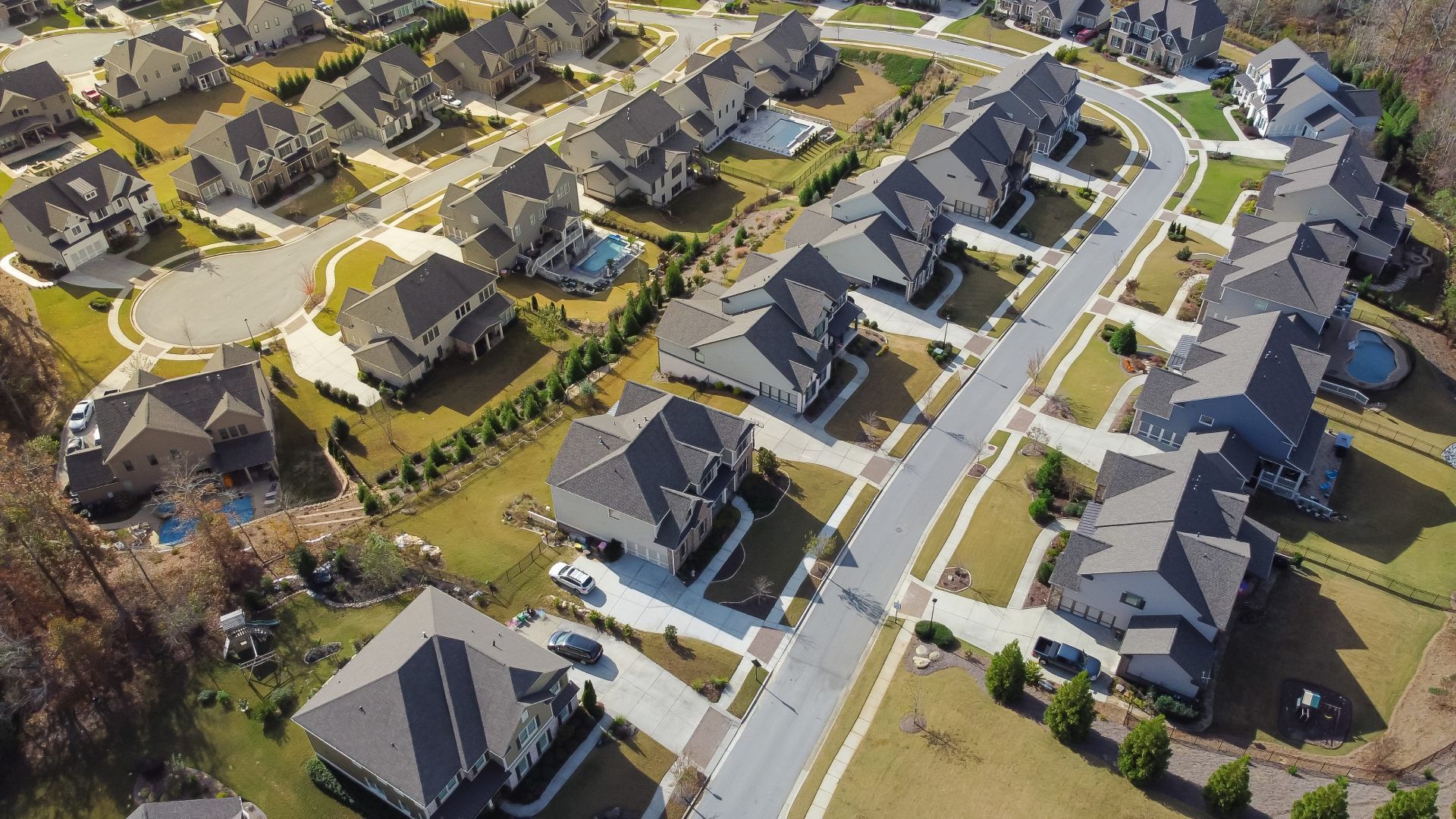 Aerial view of a suburban neighborhood with rows of houses, roads, and green lawns, some with pools.