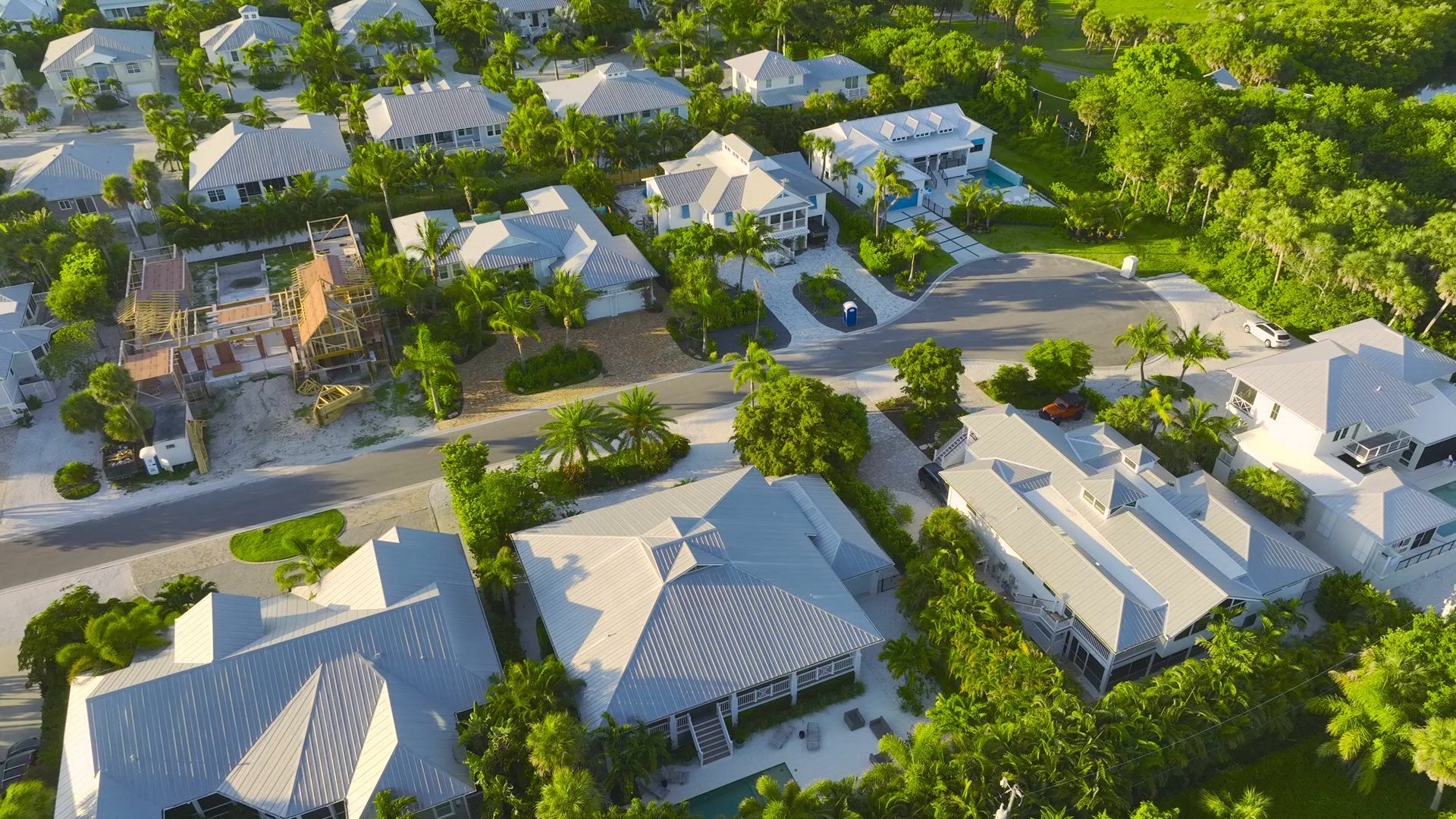 Aerial view of a neighborhood with houses and lush green trees. Gray roofs and a road are visible.