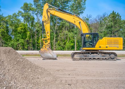 Yellow excavator parked on gravel near a pile of gravel, trees in the background.