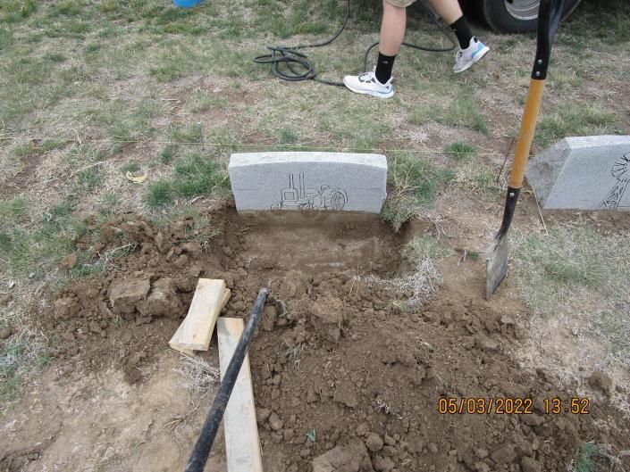 Person digging a grave beside concrete markers in a grassy cemetery.
