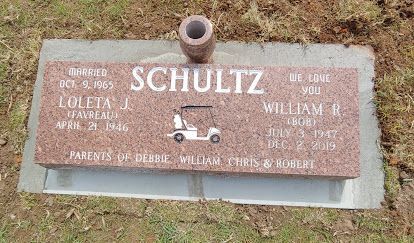 Bronze grave marker reading “SCHULTZ,” set in grass with a small bronze tractor emblem.