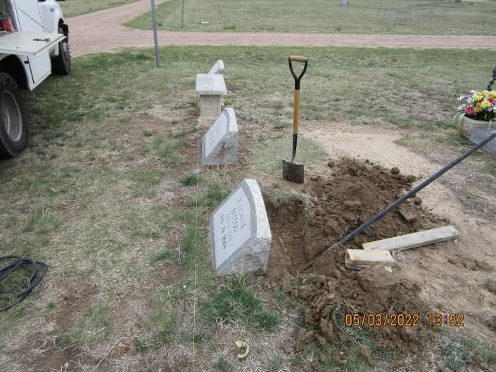 Fresh grave site with white headstones, a shovel, and a dirt hole beside a parked truck on grass