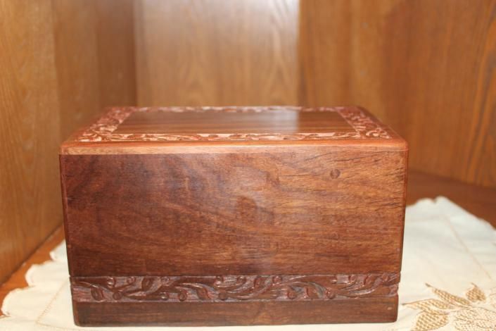 Small carved wooden box with a glossy lid, sitting on a light cloth against a wood-paneled background