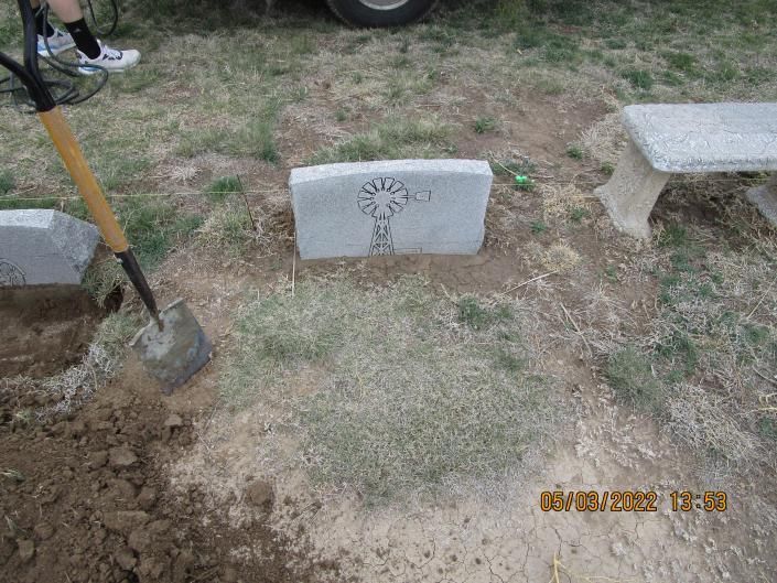 Grassy plot with a small concrete marker, shovel at left, and bench at right