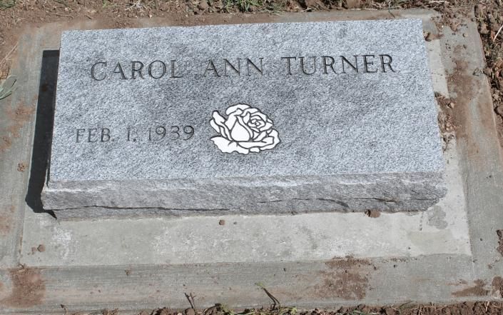 Gray granite gravestone engraved “CAROL ANN TURNER” with a white rose emblem and date “FEB. 1, 1949.”