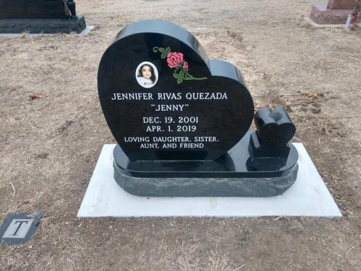 Black heart-shaped gravestone with flowers and a small angel, set on a white base in a cemetery.