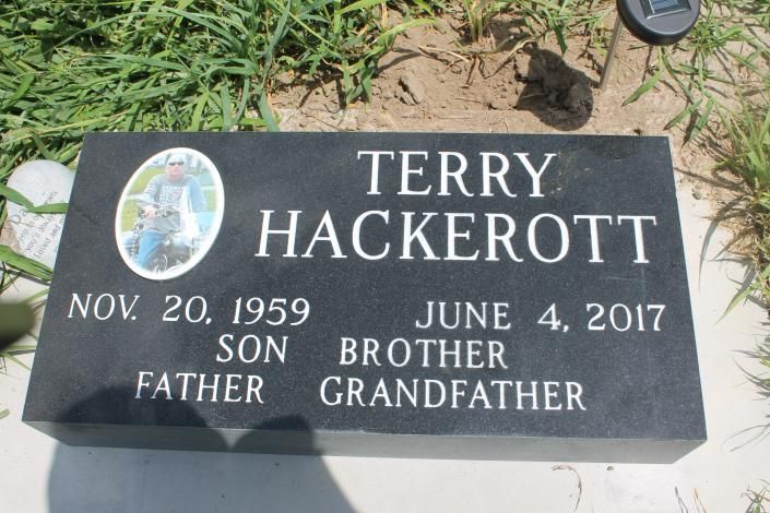 Black gravestone for Terry Hackerott with dates and a small oval photo, set in grass.