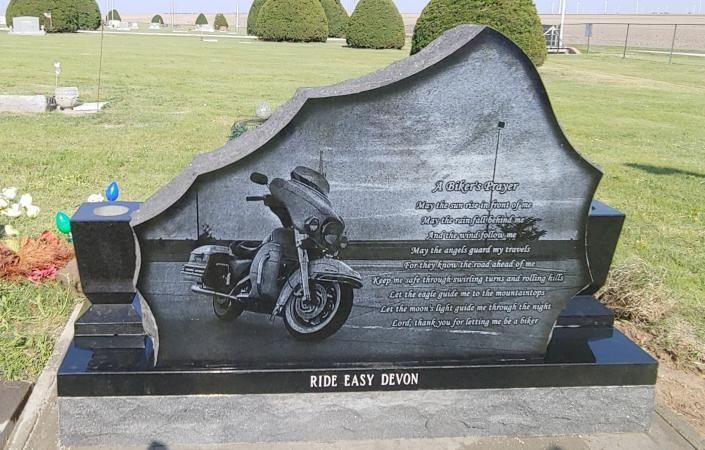 Black motorcycle memorial headstone in a cemetery with engraved text and flowers on a small vase