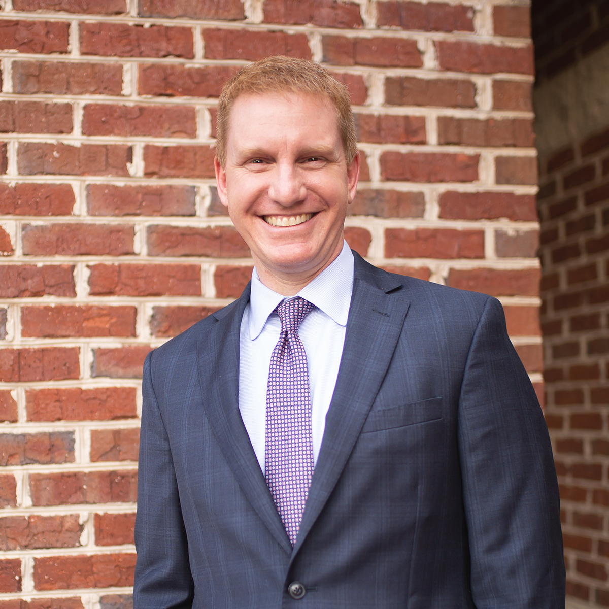 Man in suit smiles in front of a brick wall, wearing a purple tie.