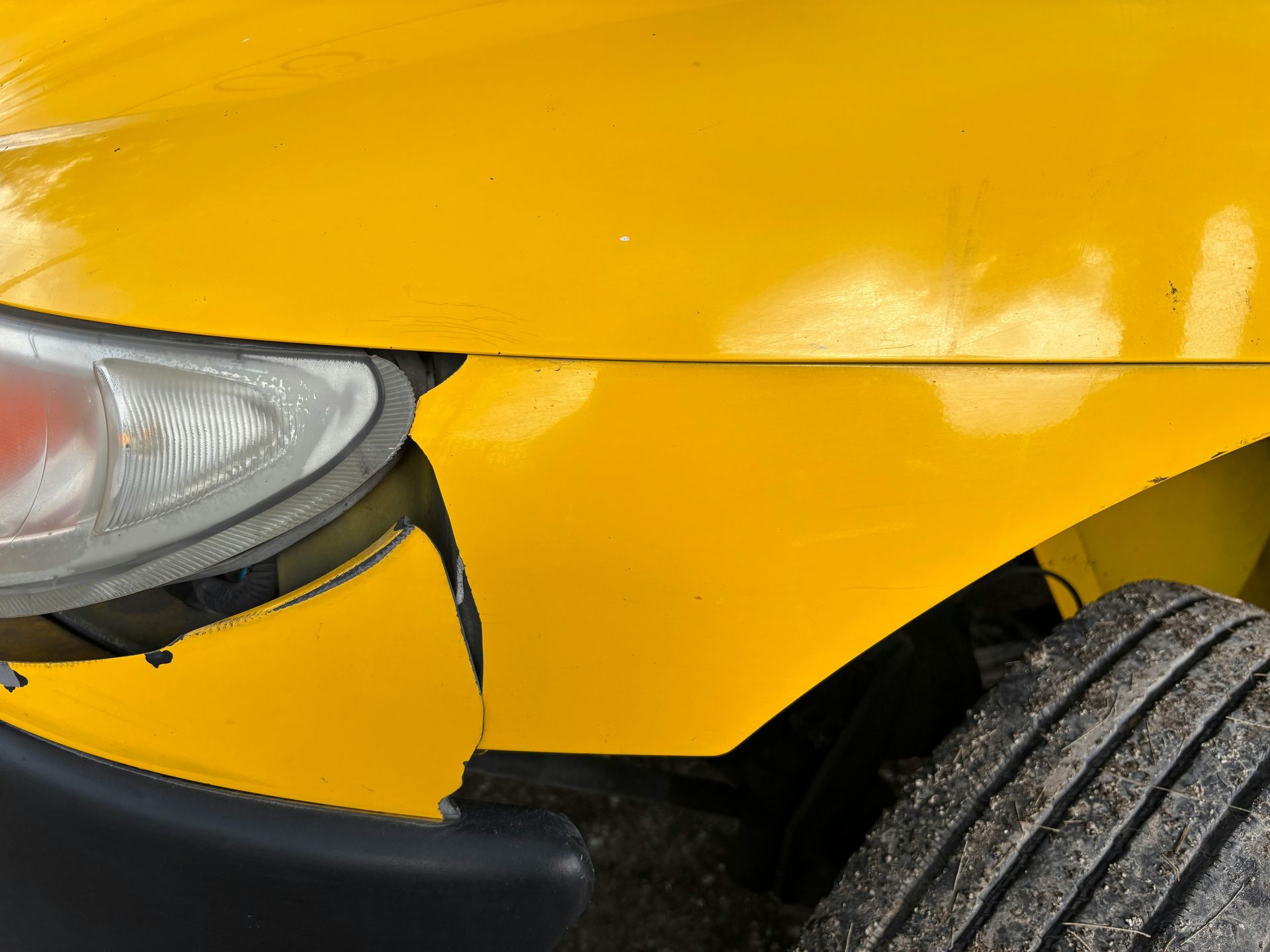 Yellow car with a damaged front bumper and headlight, next to a tire.