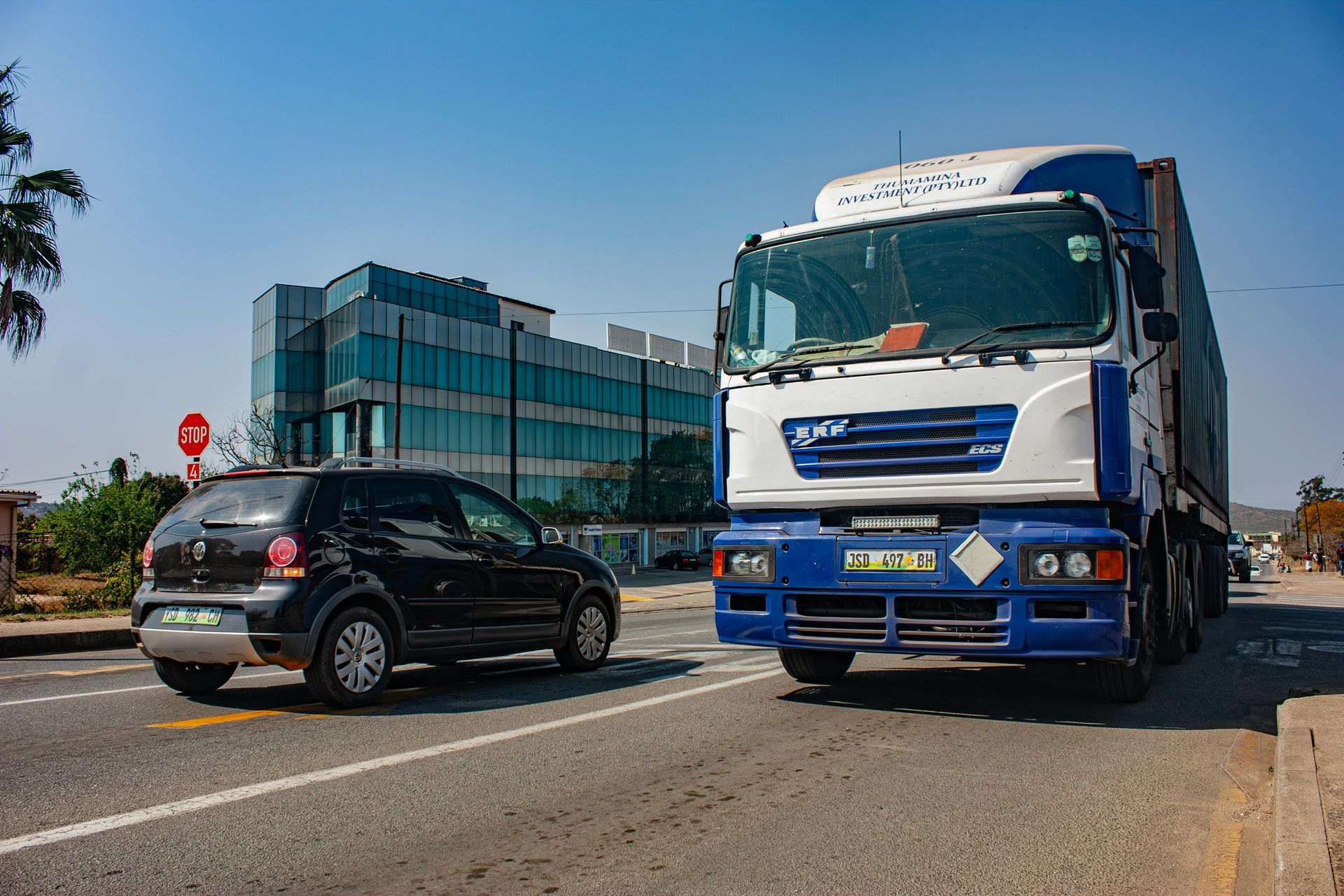 A black car and a large truck on a road in front of a modern building under a blue sky.