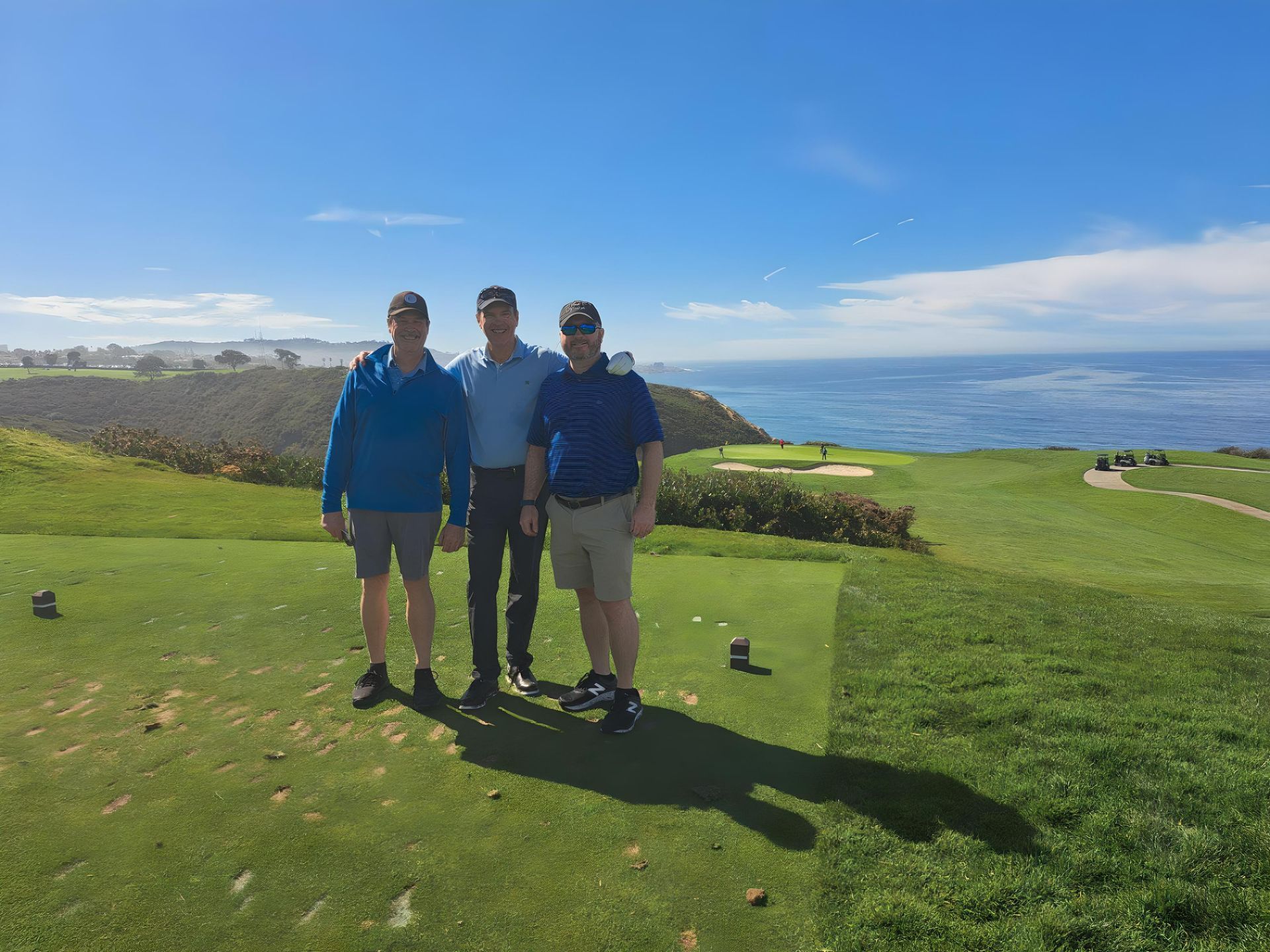Three men are standing on a golf course with the ocean in the background