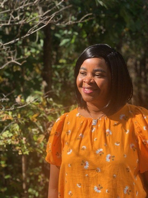 A woman in an orange shirt is standing in front of a forest.