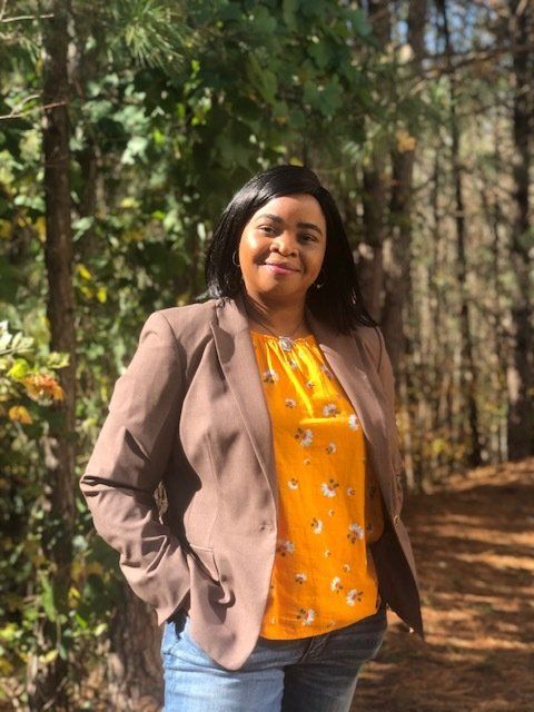 A woman in a brown jacket and a yellow shirt is standing in the woods.